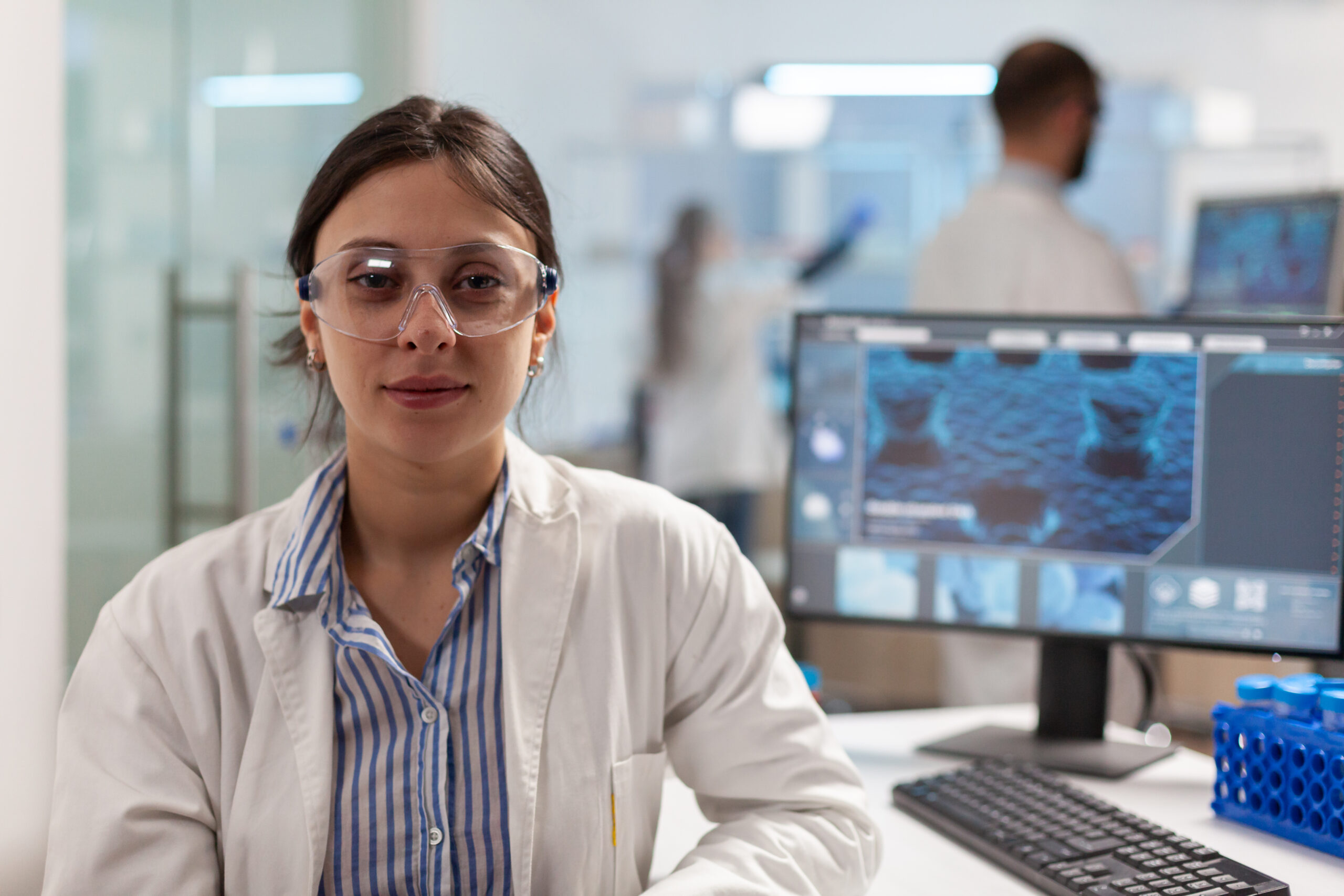 Scientist with lab coat sitting in laboratory looking at camera smiling, while colleague working on computer in background. Chemist examining virus evolution using high tech for scientific research, vaccine.